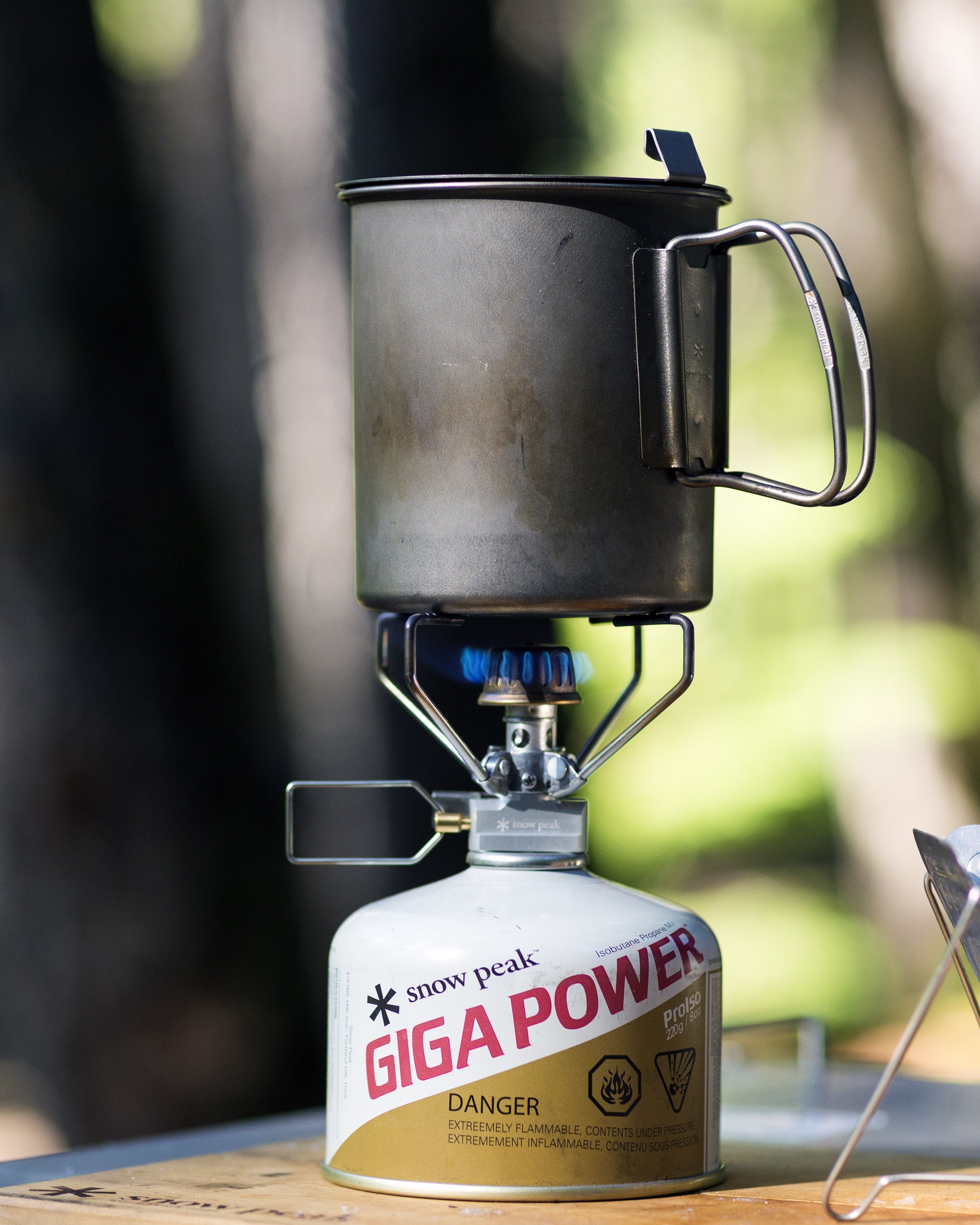 A portable camping stove, the Snow Peak GigaPower, is being used to heat water in a cup outdoors. The stove is fueled by a canister and has a warning label about flammable contents.