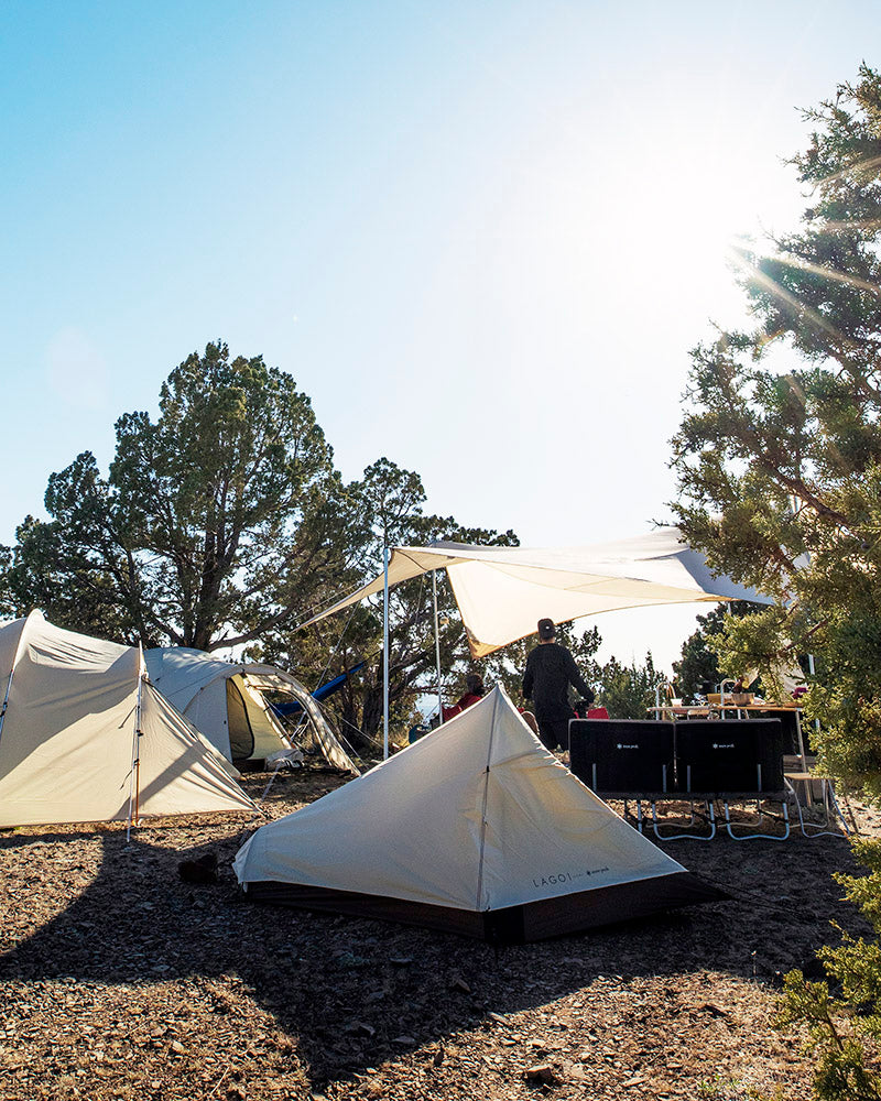 A camping setup with several tents and a tarp providing shade. The tarp is supported by poles and is stretched over a camping table and chairs.